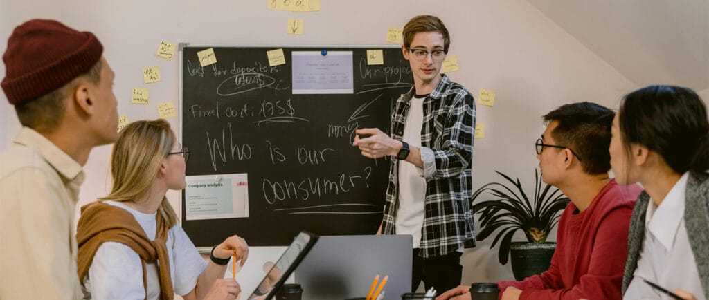 Group of attendees on a Spontaneous Think Tank workshop, brainstorming over a wall of sticky notes and a blackboard while they listen to the workshop leader.