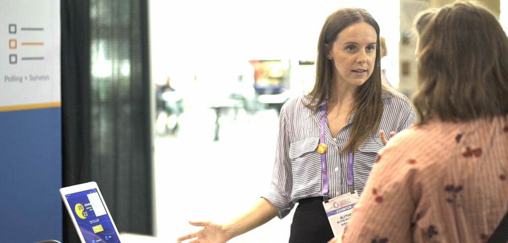 A woman in a striped shirt and wearing an EventMobi badge has a conversation while gesturing to an iPad. 