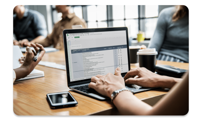 Mehrere Menschen um einen Schreibtisch herum, im Vordergrund jemand bei der Arbeit an einem Laptop, auf dem die kundenspezifisch individualisierte Checkliste von EventMobi geöffnet ist.